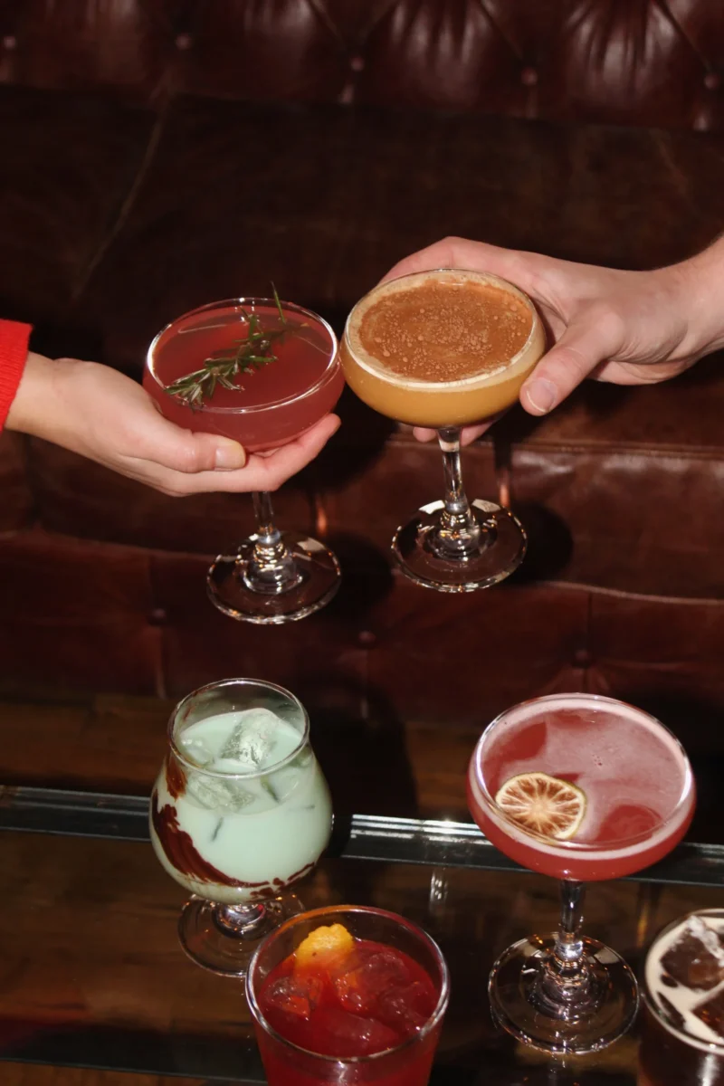 A variety of cocktails sit on a wood table as two hands each grab one.