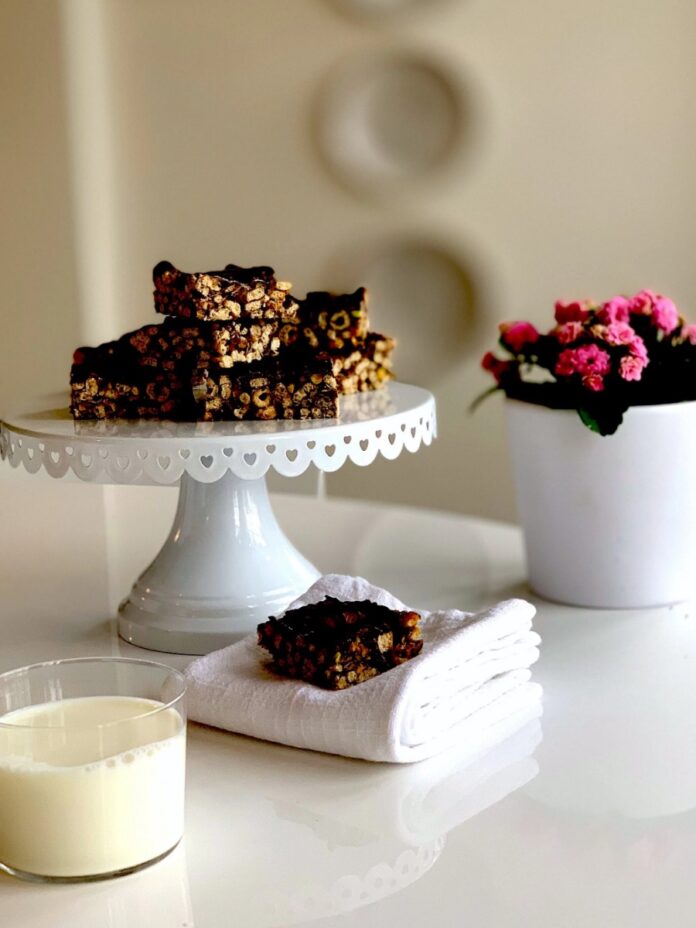 A vertical stack of dark, chocolate-covered no-bake Cheerio bars on a white scalloped cake stand, with a single bar on a white napkin and a glass of milk next to a potted pink flowering plant.