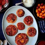 Seven mini pancake pizzas topped with tomato sauce, melted cheese, and halved cherry tomatoes on a light gray oval platter, surrounded by bowls of pepperoni slices, parmesan cheese, and grape tomatoes.