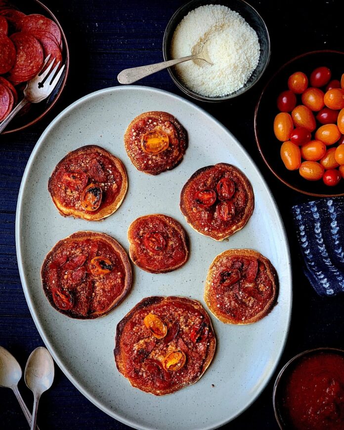 Seven mini pancake pizzas topped with tomato sauce, melted cheese, and halved cherry tomatoes on a light gray oval platter, surrounded by bowls of pepperoni slices, parmesan cheese, and grape tomatoes.