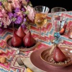 Three poached pears in a red color sit on a small plate while a larger plate and bowl holds another full pear off to the side of the table setting.