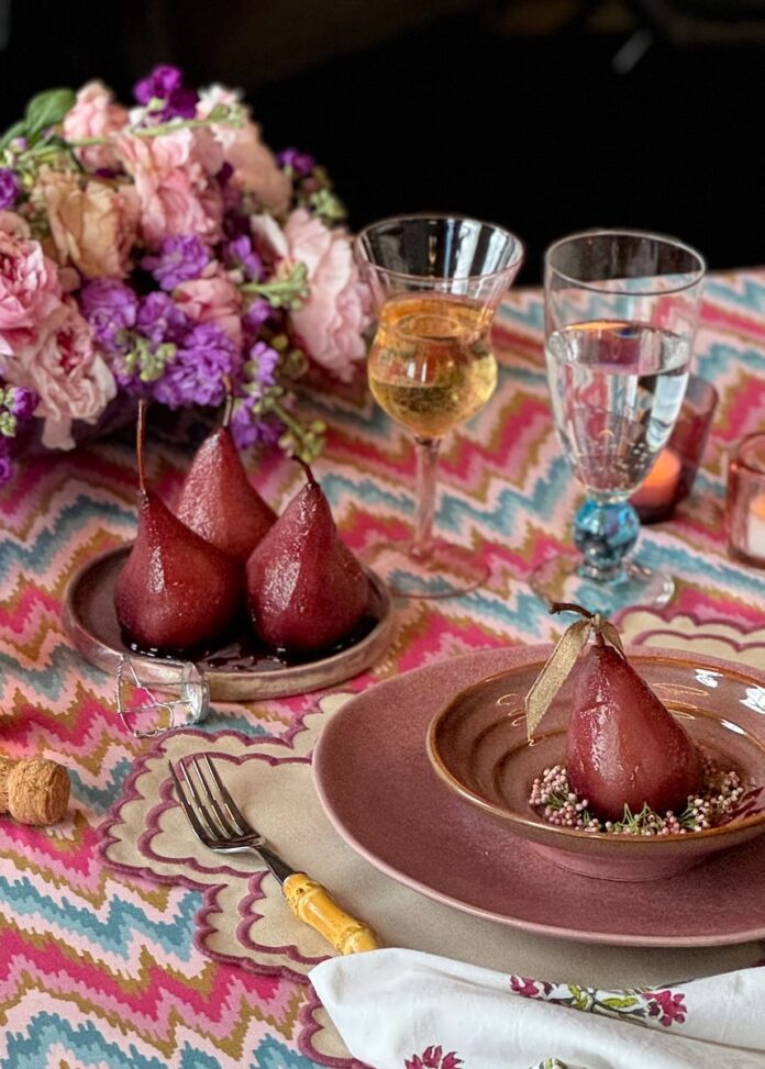 Poached Pears in Spiced Red Wine TABLE Magazine Three poached pears in a red color sit on a small plate while a larger plate and bowl holds another full pear off to the side of the table setting.