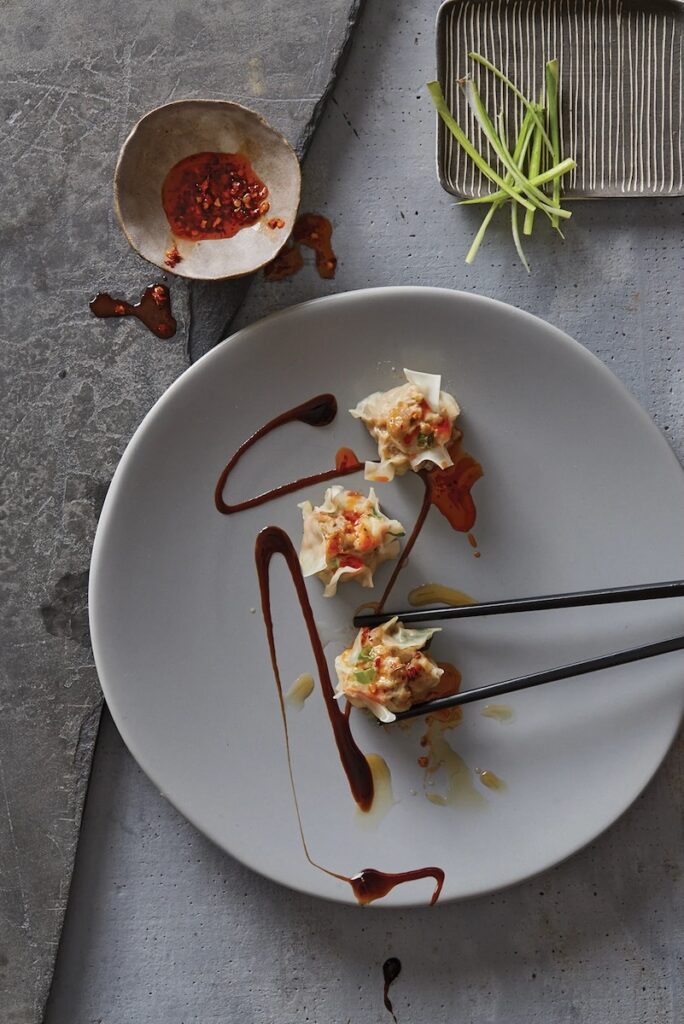 A plate of Pork and Shrimp Shumai dumplings hold with chopsticks with a sauce drizzled over top.
