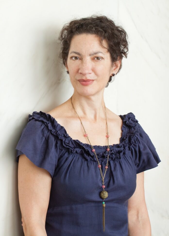 Quantum Theatre Karla Boos Retire TABLE Magazine Karla Boos of Quantum Theatre stands in a blue blouse and gold necklace against a white wall.