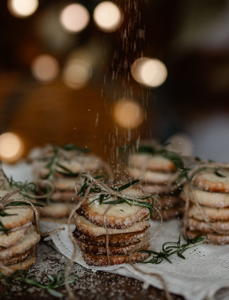 Stacks of Rosemary Butter Cookies sit wrapped in twin while someone sprinkles sugar overtop.