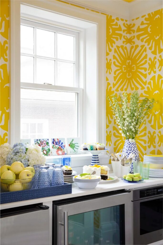 A bright and colorful kitchen nook featuring a white window, yellow and white patterned wallpaper, and a counter with a mini-fridge, showcasing desserts, glasses, and a vase of white flowers.