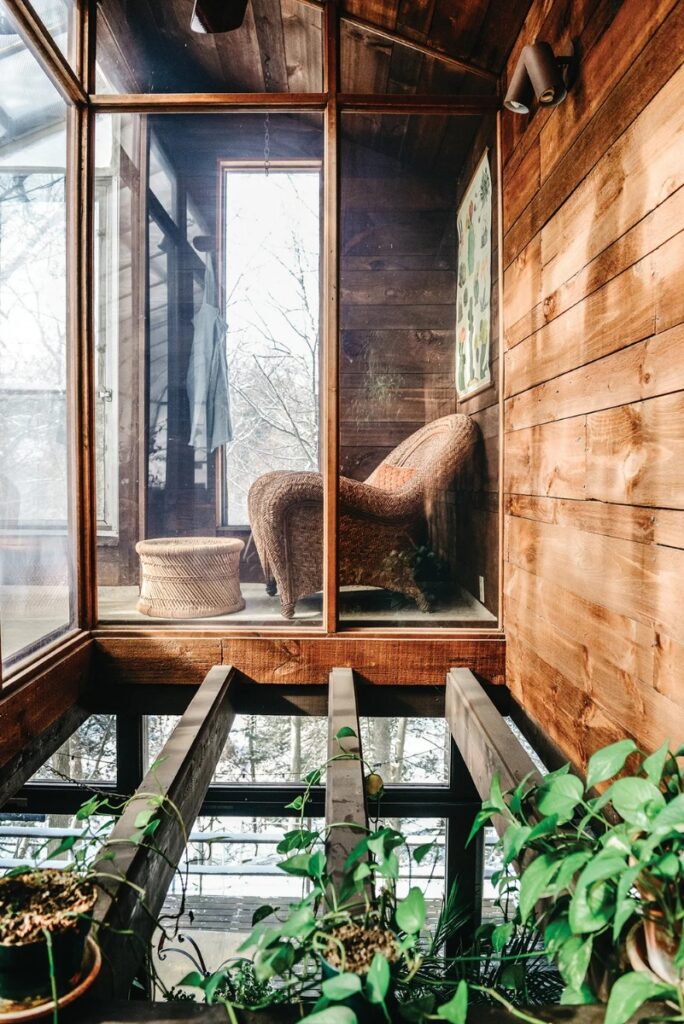 A two-story greenhouse with wood-paneled walls, featuring a woven rattan chair and stool inside a glass enclosure, with houseplants on the wooden floor beams in the foreground.