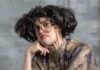 Treasure Treasure is ‘Agnus Teaches Acting’ A woman in a dress leans against a chair with a frown on her face, crazy brown hair, and huge glasses.
