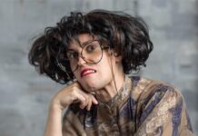 Treasure Treasure is ‘Agnus Teaches Acting’ A woman in a dress leans against a chair with a frown on her face, crazy brown hair, and huge glasses.