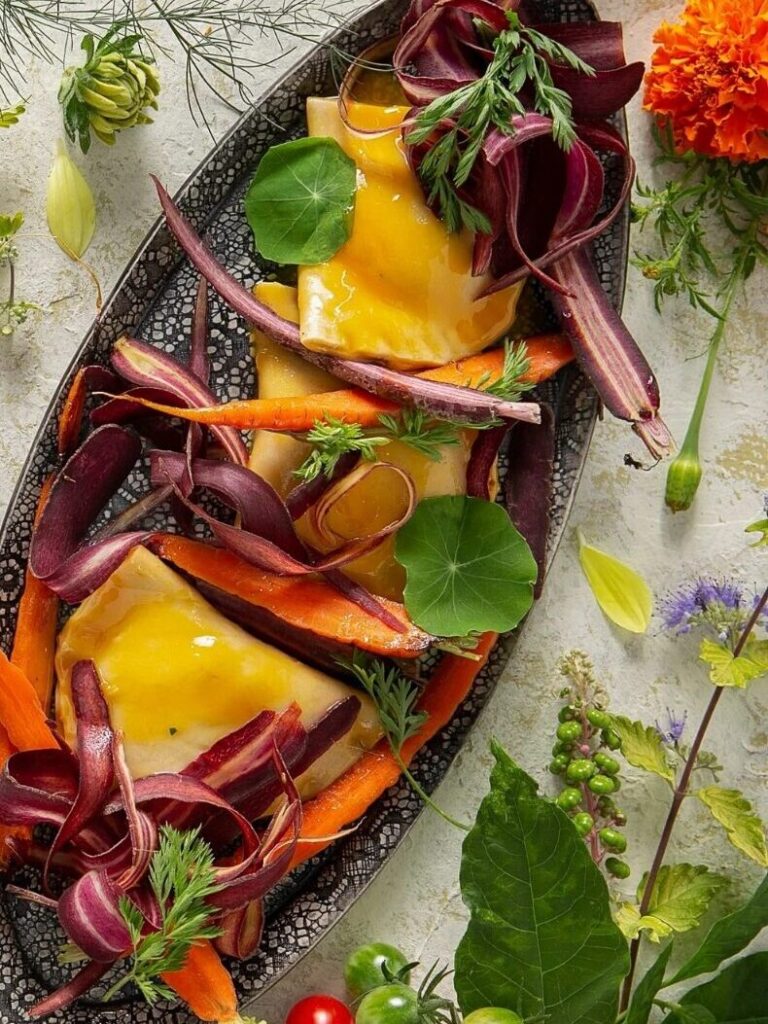 A plate of carrot pierogies garnished with carrot ribbons and fresh herbs, surrounded by flowers and green and red vegetables.