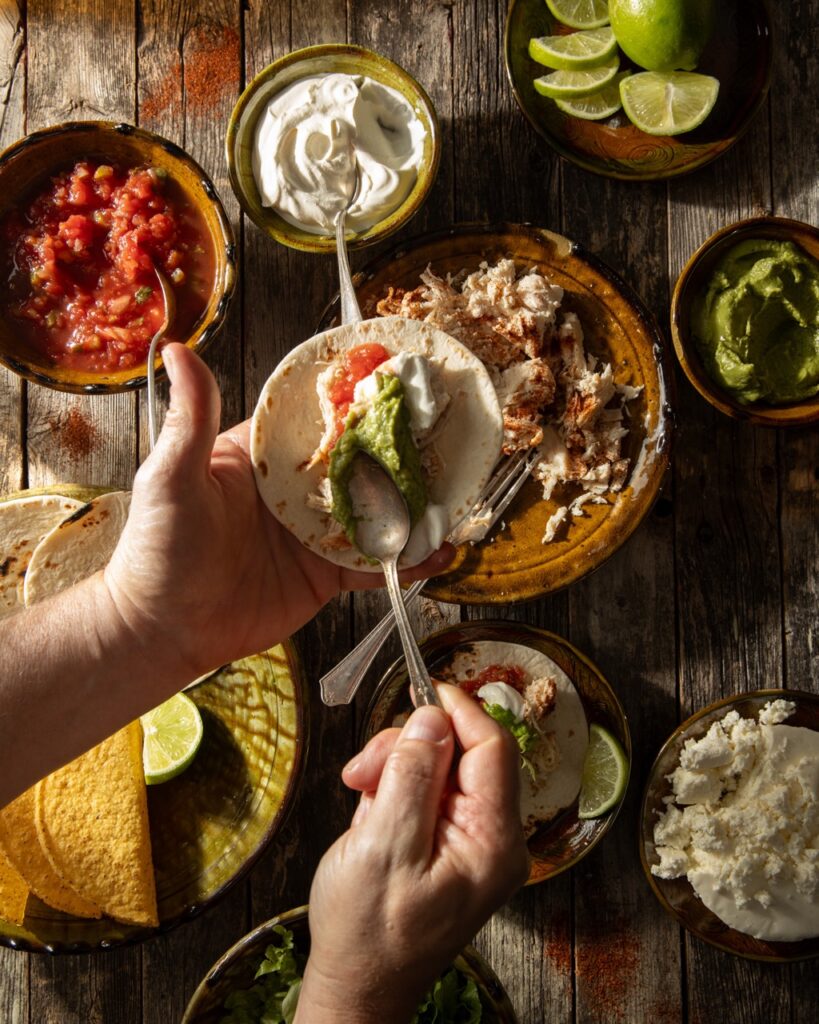 A person smears guac on a taco shell over a table of taco toppings.