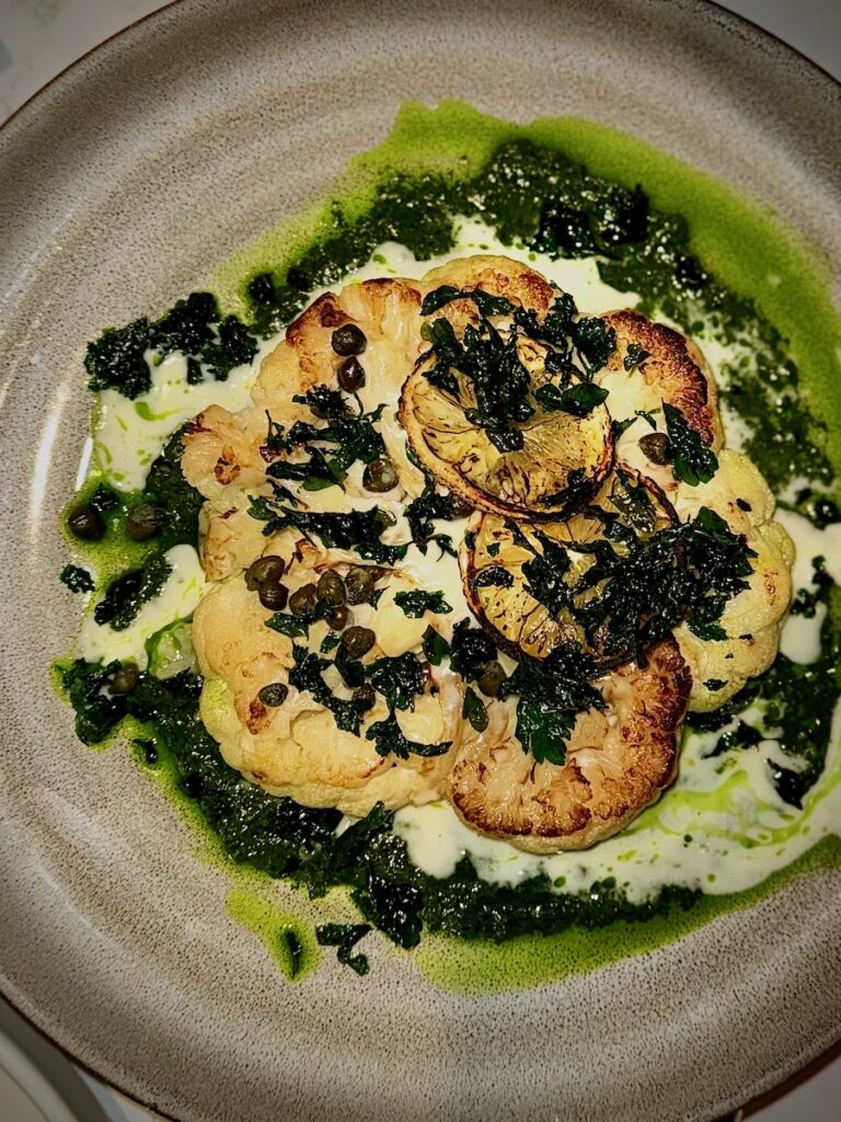 A cauliflower steak sits in a basil and lemon sauce with capers and lemons on top.