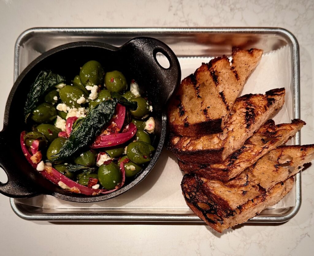 A pan of olives sits beside slices of bread.