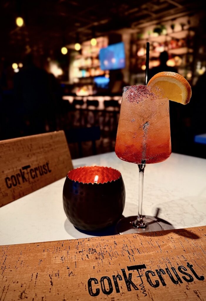 An orange cocktail sits beside a candle and Cork & Cork menu on a white table.