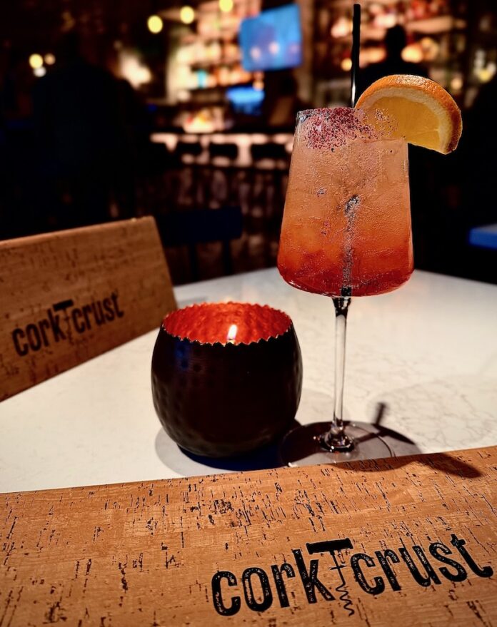 An orange cocktail sits beside a candle and Cork & Cork menu on a white table.