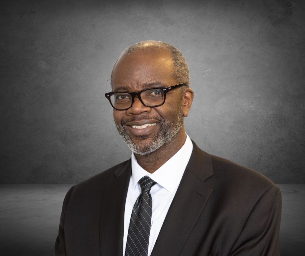 John Wallace sits in a black suit and tie against a black background.