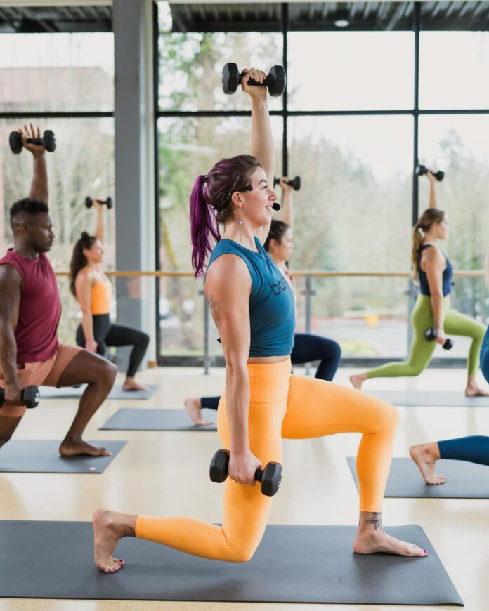 A woman coaches a fitness class while lifting weights and lunging.