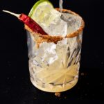 A liquid pours into a glass with a tajin rim, lime slice, and hot pepper.
