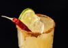 Hot Bunny Rum Smash (A Bad Bunny Cocktail) A Hot Bunny Smash in a yellow color sits in a cocktail glass with a tajin rim, lime slice, and hot pepper garnish.