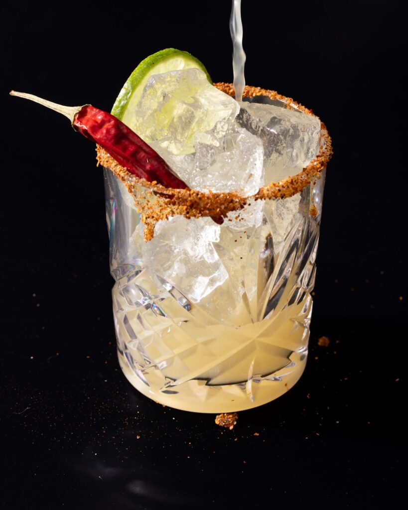 A liquid pours into a glass with a tajin rim, lime slice, and hot pepper.