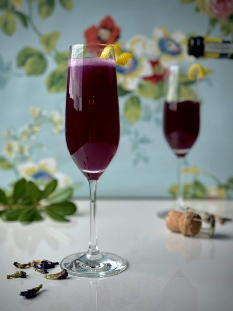 Two champagne flutes of a dark purple Lord & Lady Bridgerton cocktail with a lemon peel twist garnish, butterfly pea flowers below, and a bottle of champagne pouring into the background glass. It's all set against a blue floral background.