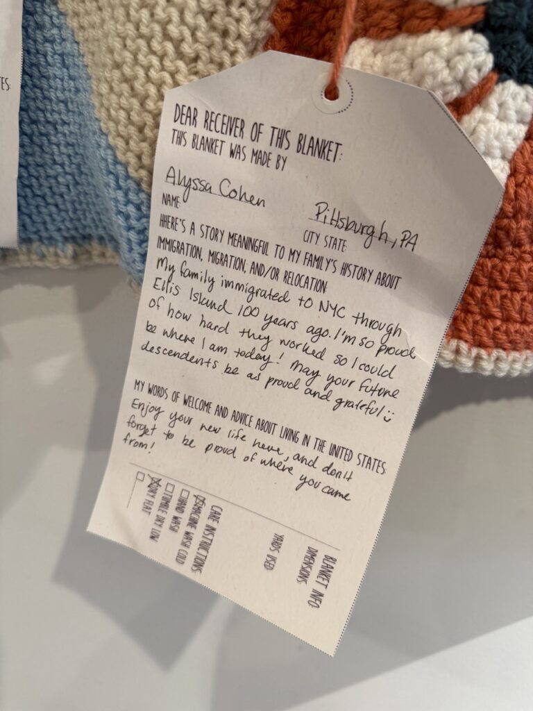 A white tag hangs from a knit object on a wall.