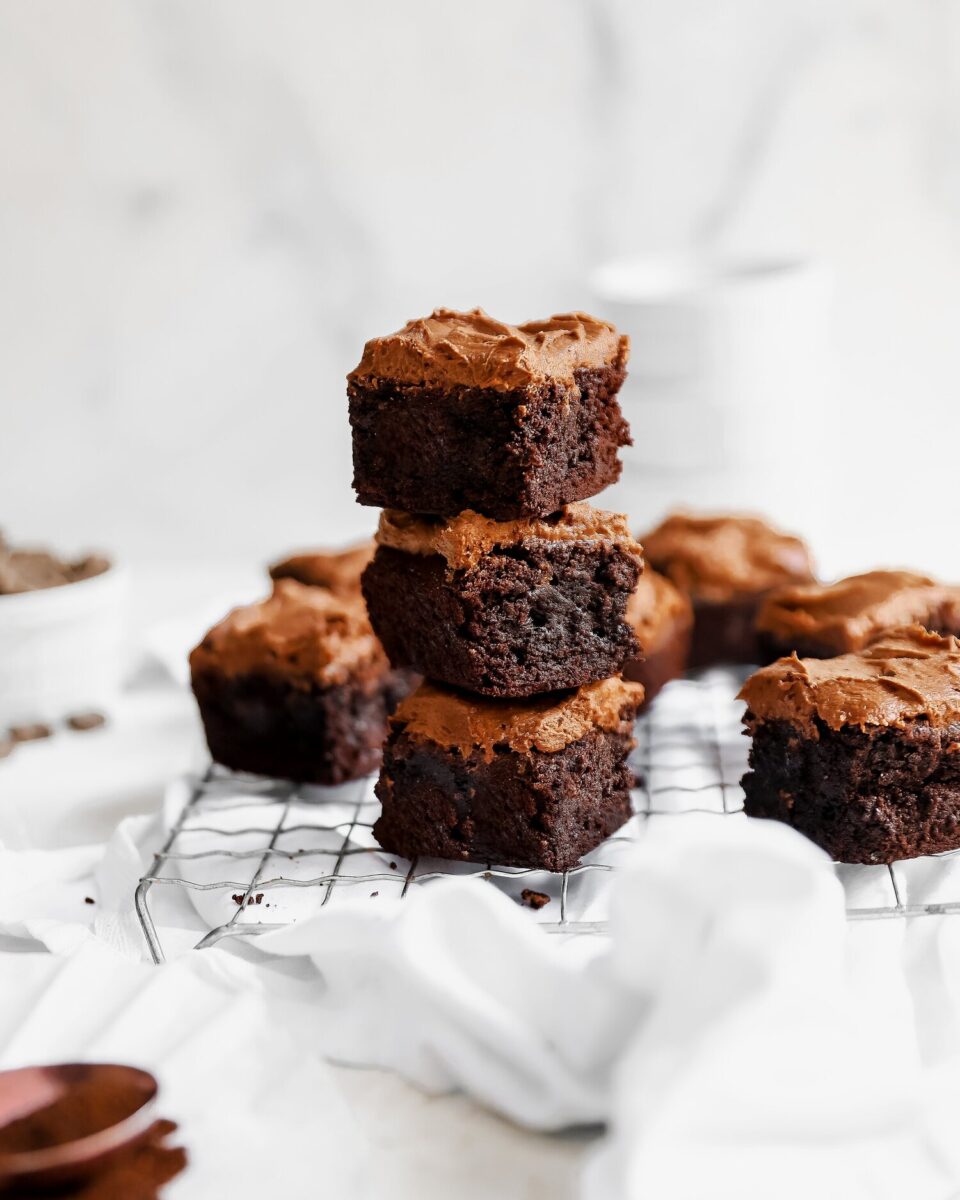 A stack of brownies with a mocha chocolate frosting sits on a cooling rack amongst white sheets.