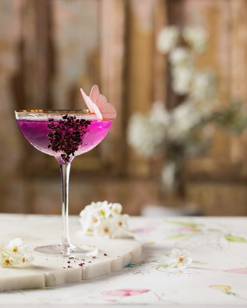 A coupe glass with a purple Mother's Day Gin and Tonic mocktail with crushed freeze-dried blueberries on the side and an edible pink butterfly on the rim. White flowers are scattered nearby.