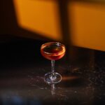 A cocktail in am amber color sits in a tall glass on a table in the evening light.