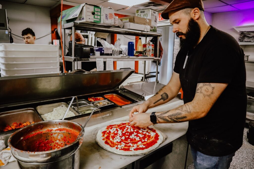 A man puts toppings on to a pizza at a station.