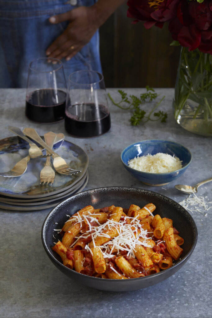 Stanley Tucci Inspired Pasta alla Norma sits in a bowl to the right with two glasses of red wine off to the back left.