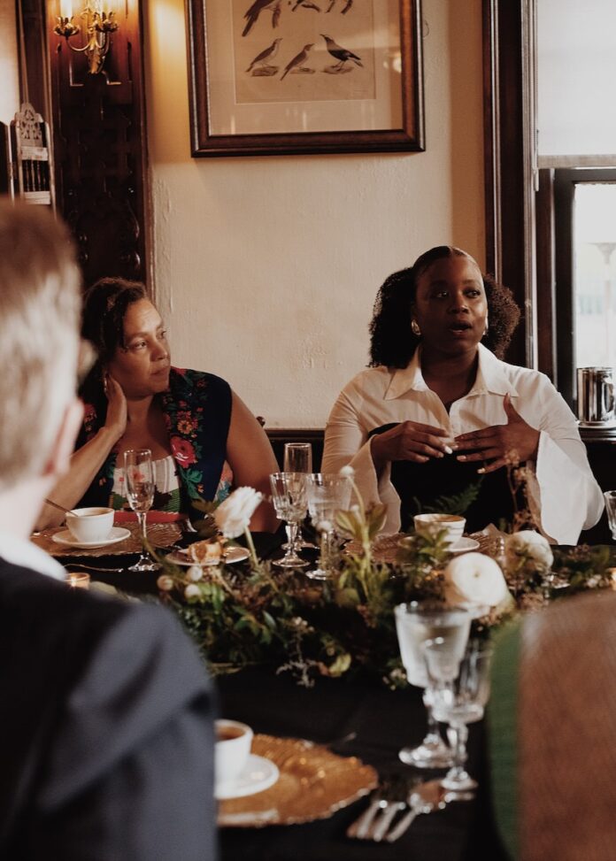 A woman in white and black sits at a table talking to the people across from her.