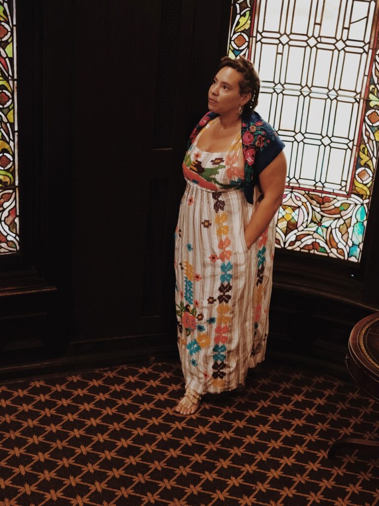 A woman stands in a long white dress with colors all over it.