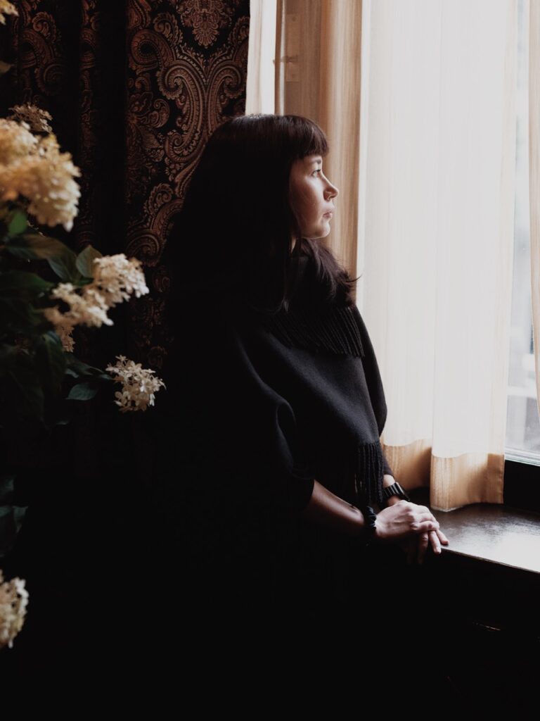 A woman looks out a window with white flowers behind her.