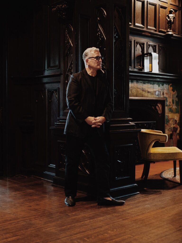 A man in black clothes leans against a dark doorway.