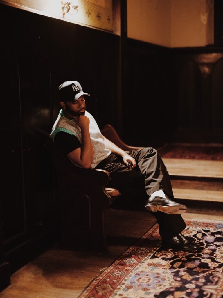A man sits in a leather arm chair with a baseball cap on.