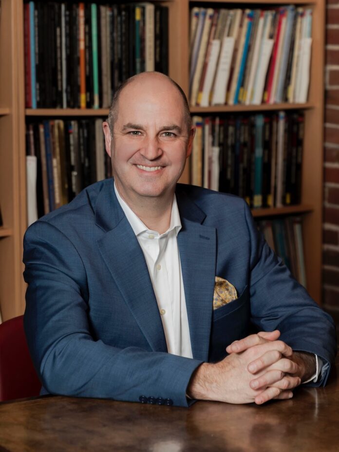 William Powers sits at a wooden desk in a blue suit jacket and white dress shirt.