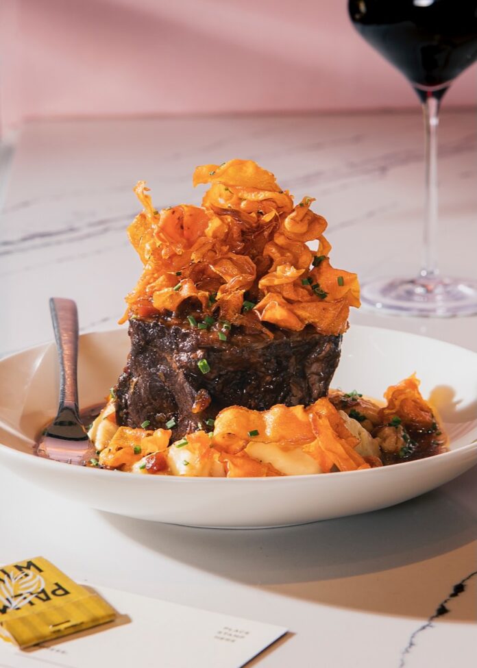 A white plate with a chunk of steak and pasta over top of it all on a marble table with a glass of wine in the background.