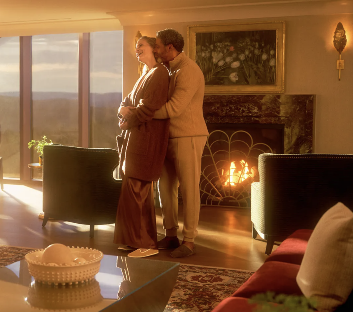 A couple stands in a Valentine's Day Hotel package suite at Nemacolin with a fireplace in the distance as they look out the long windows.