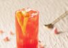 Rom-Com Cocktails and Movie Pairings for Spicing Up Your Valentine’s Day In a tall glass sits a red Sweet Home Alabama Slammer garnished with a heart formed orange peel and surrounded by pink roses on the table.