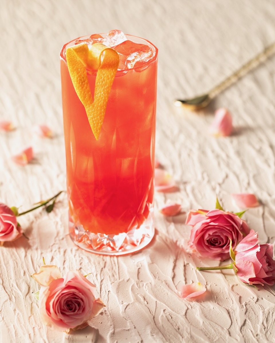 In a tall glass sits a red Sweet Home Alabama Slammer garnished with a heart formed orange peel and surrounded by pink roses on the table.