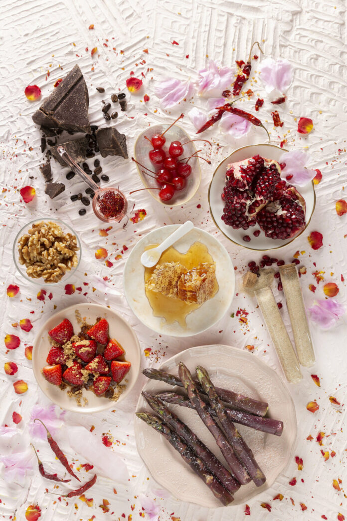 An overhead photo of aphrodisiac foods like cherries, chocolate, asparagus, walnuts, honey, strawberries, chilies, saffron, and pomegranate in small bowls and on little dishes on a textured white surface with rose petals.