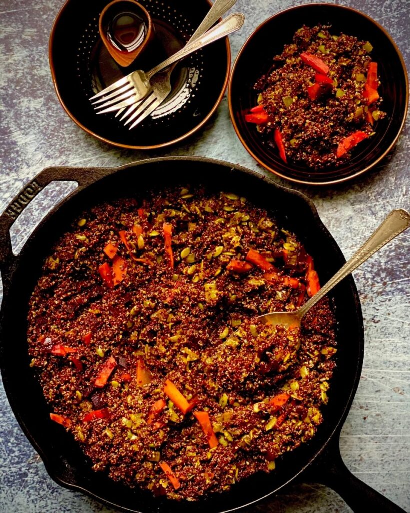 A cast iron skillet with toasted red quinoa and pickled vegetables, with two black bowls and a fork on the side.