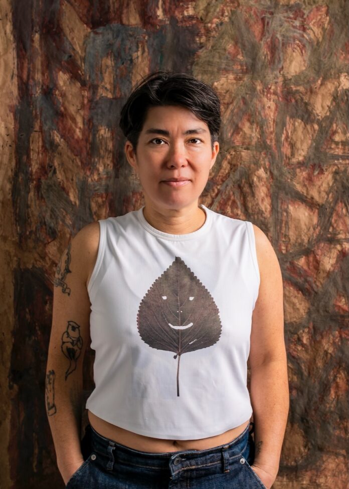 A woman with short black hair in a white muscle top stands in front of a wood backdrop.