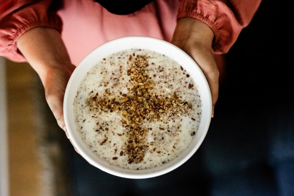 Hands holding a white bowl of Sheerbrinj, a traditional Afghan sweet rice pudding garnished with cardamom and crushed almonds.