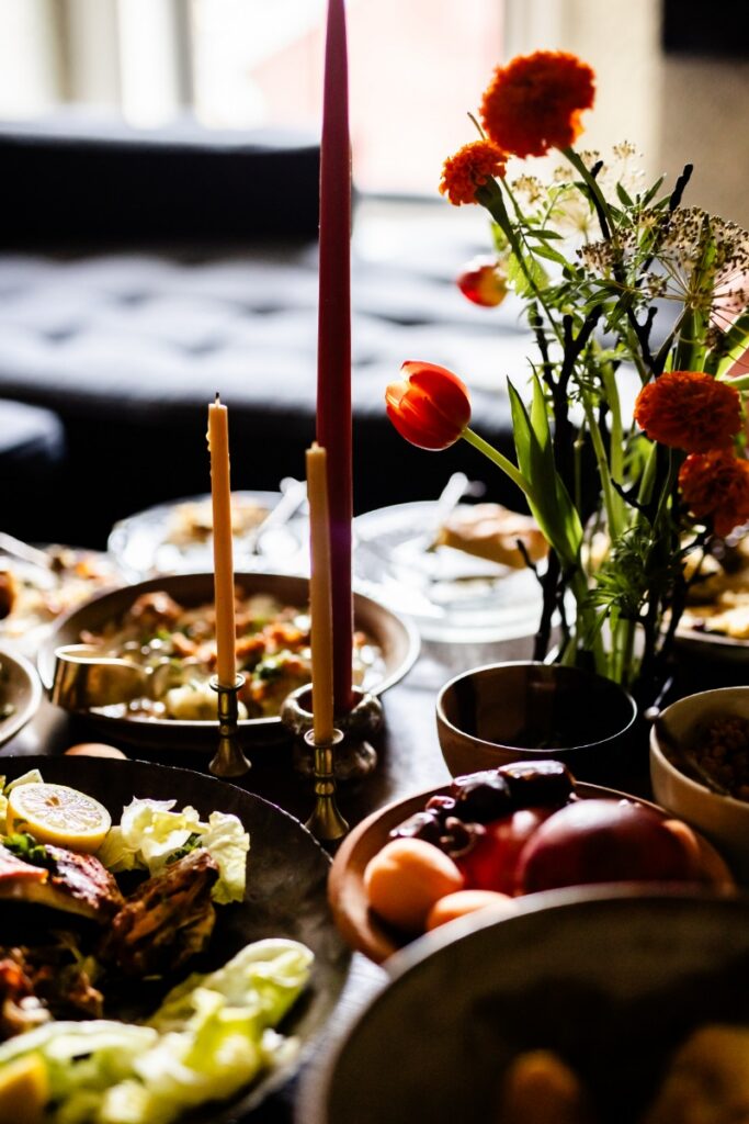 A festive table setting featuring tall taper candles, vibrant orange flowers, and a traditional Afghan meal including grilled meats and bowls of fresh fruit.