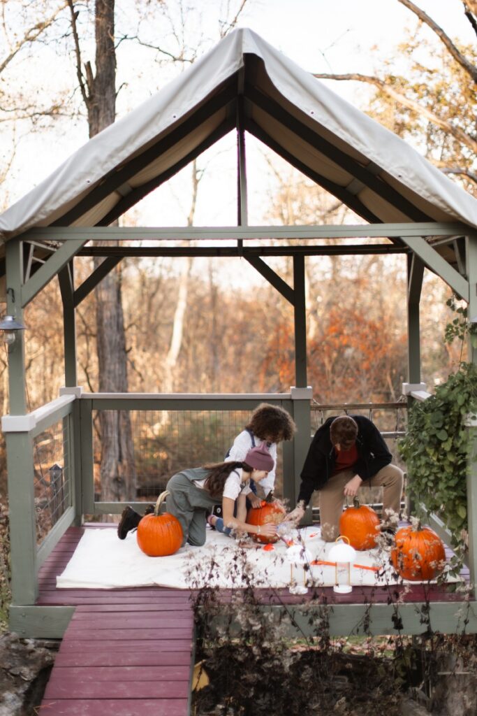 A group of children gathered inside a covered outdoor play "fort" on stilts, gutting and carving large orange pumpkins on a white sheet during a fall gathering.
