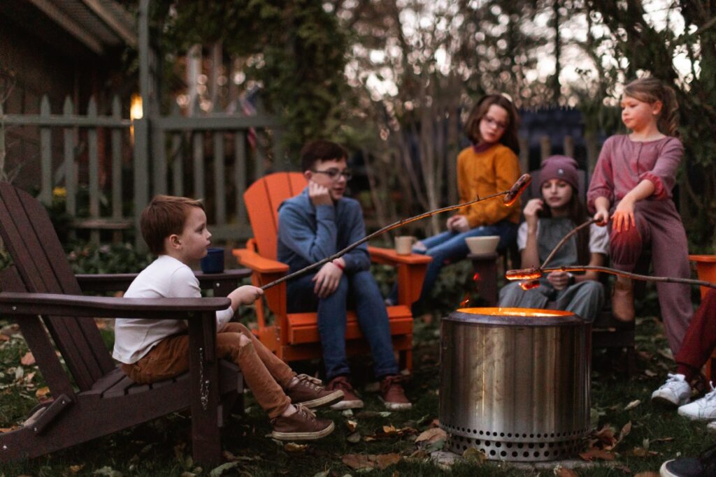 A group of children sitting in Adirondack chairs roasting sausages on sticks over a modern silver fire pit during a backyard autumn gathering.