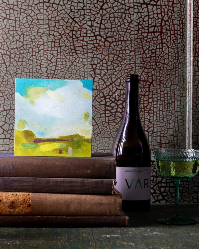 An intimate landscape painting by April Ryan resting on a stack of vintage books next to a bottle of Vara wine and a green glass of champagne.