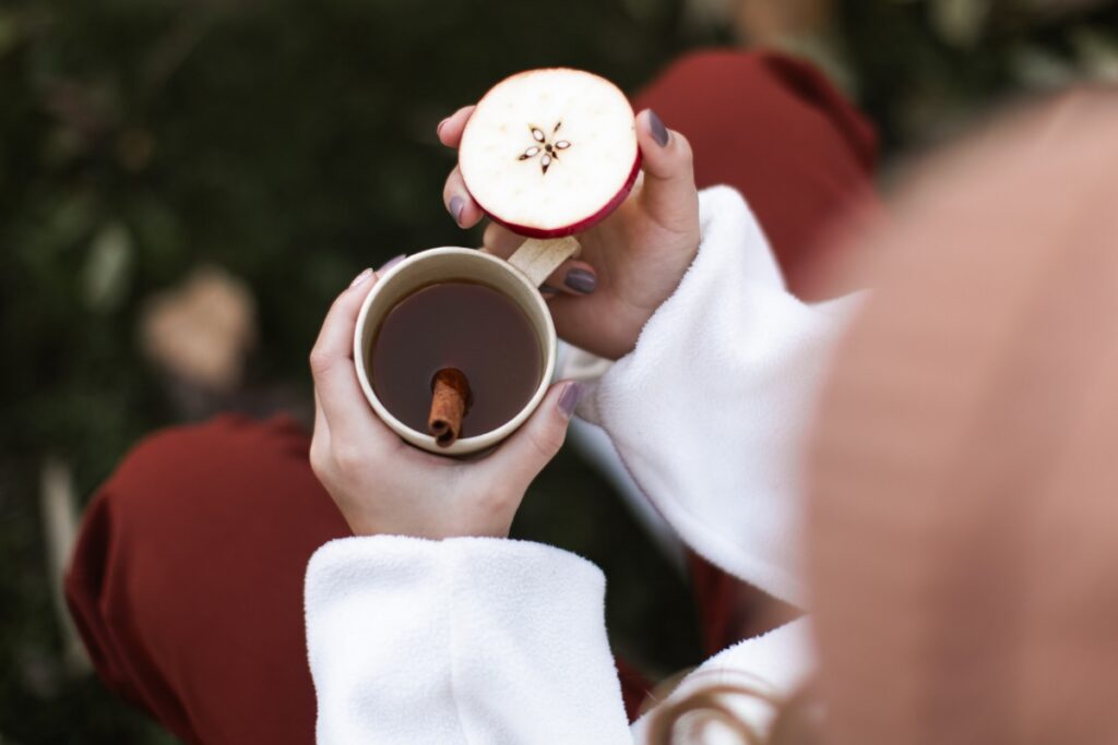 A person holding a mug of warm apple cider with a cinnamon stick and a fresh apple slice during a fall gathering.